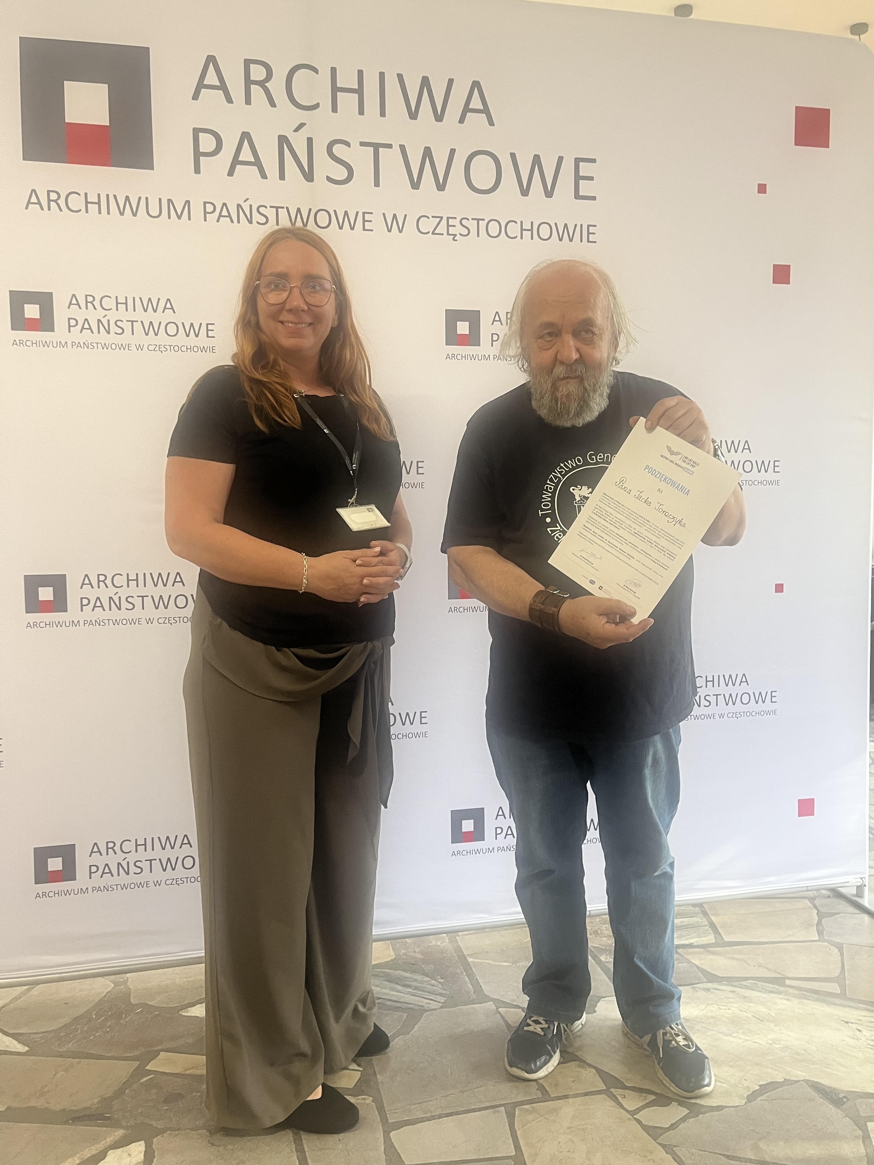 Kobieta i mężczyzna stoją na tle banneru reklamowego Archiwum - mężczyzna trzyma w rękach dyplom